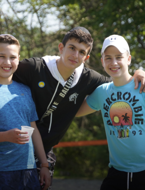 A group of children engages in a playful struggle, with one child being gently pulled by two peers, against a lush outdoor backdrop.