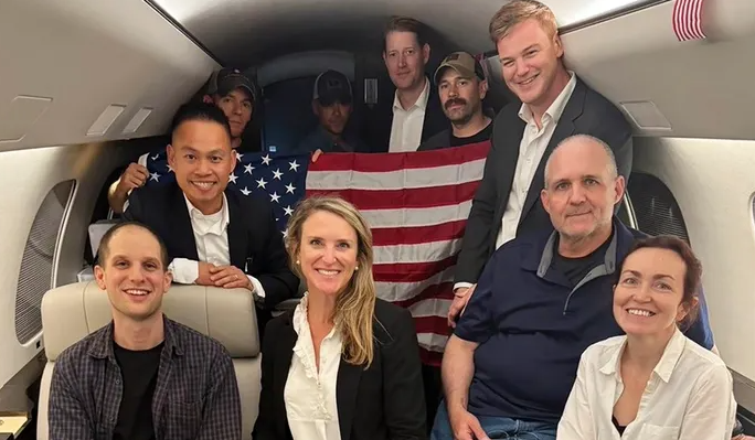 A group of people in a jet cabin, some holding a U.S. flag, smiling at the camera.