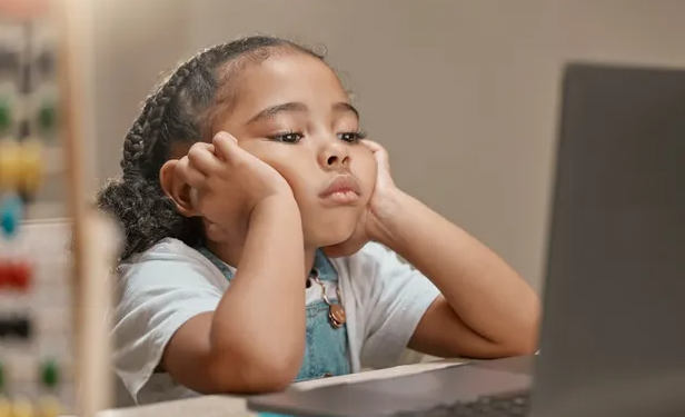 A young girl sits at a desk, resting her hands on her head, appearing deep in thought or contemplation.