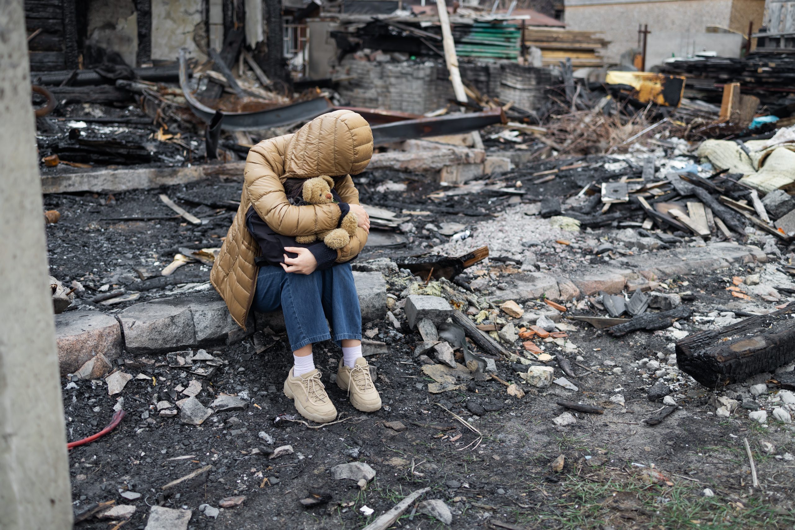 Young woman sits on ground with head on knees in aftermath of natural disaster.
