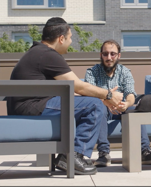 Two men sit together on a rooftop patio, engaged in a conversation, surrounded by greenery and comfortable seating.
