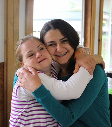 Two young woman smile and embrace in a camp bunk.